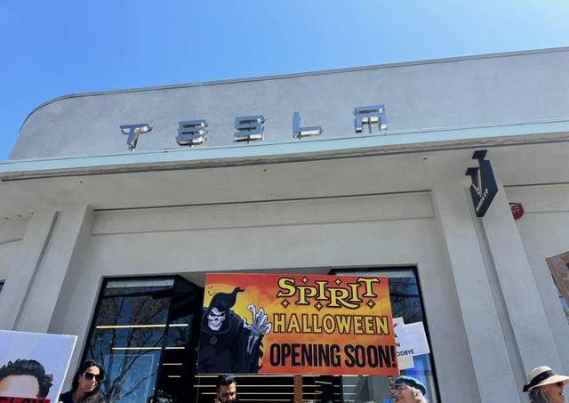 Protester sign in front of a Tesla dealership that shows an ad for Spirit Halloween store coming soon